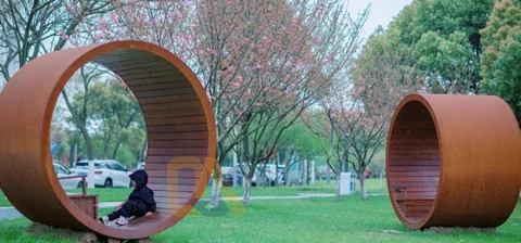 Two cylindrical seating shelters on a lawn beneath trees.